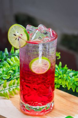Kırmızı gazozu buzla ve limon dilimleriyle tazeleyerek berrak bir bardakta servis edilir, arka planda yeşil yapraklarla ahşap bir yüzeye yerleştirilir..