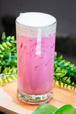 Buzlu pembe süt, kremalı, uzun bir bardakta servis edilir, buzla doldurulur ve arka planda yeşil yapraklar bulunan ahşap bir yüzeye yerleştirilir..