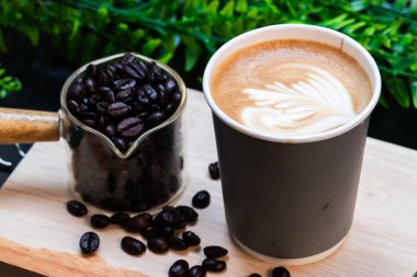 Tahta bir tahtanın üzerinde kavrulmuş kahve çekirdeği konteynırının yanında servis edilen, kalp latte sanatıyla dolu kağıt bir fincan kahve..