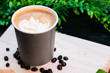 Tahta tahtada güzel latte sanatıyla kaplı, dağılmış kahve çekirdekleri ile çevrili kağıt bardak sıcak latte..