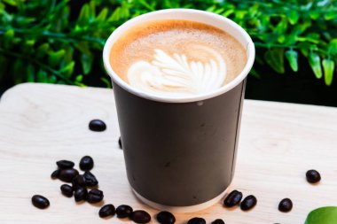 Tahta tahtada güzel latte sanatıyla kaplı, dağılmış kahve çekirdekleri ile çevrili kağıt bardak sıcak latte..