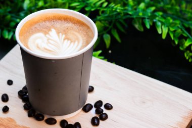 Tahta tahtada güzel latte sanatıyla kaplı, dağılmış kahve çekirdekleri ile çevrili kağıt bardak sıcak latte..