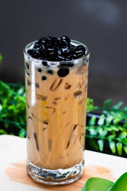 Buzlu sütlü çaya yakın plan, siyah tapyoka incileri uzun bardakta, ferahlatıcı köpüklü çay, ahşap masa üzerinde yeşil yapraklı arka plan..