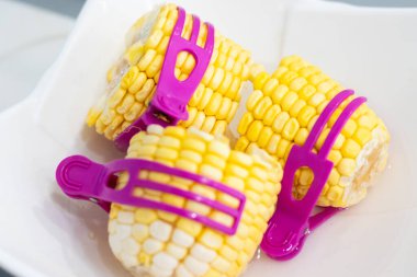 Beyaz bir tepside servis edilen, parlak mor plastik klipslerle koçandaki üç kısa sarı mısır parçasından oluşan bir yakın çekim. Mısır, sıcak bir tencerede pişirilmeye hazır..
