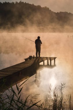 Göl kenarında güzel bir sabah sahnesi. Güneşin doğuşunda tahta bir iskelede balık tutan bir adamın silueti..