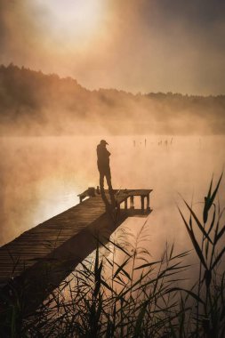 Göl kenarında güzel bir sabah sahnesi. Güneşin doğuşunda tahta bir iskelede balık tutan bir adamın silueti..