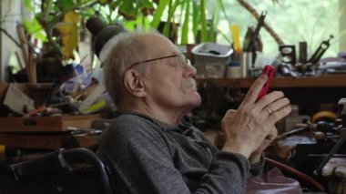 Senior elderly master jeweler in wheelchair with a smartphone in a workshop