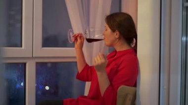 Young woman is drinking red wine while sitting at the balcony looking out the window late at night