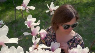 Happy woman in sunglasses enjoying nature and touching blooming magnolia flowers in the garden