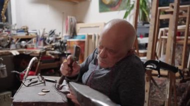 Senior elderly master jeweler in wheelchair works in a workshop with an anvil