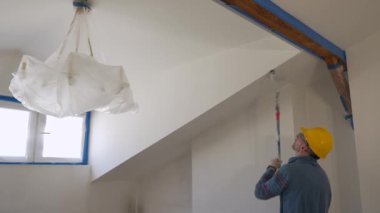 Yetişkin bir adam silindirle bir duvar boyuyor, tavan arası zemininde tamirat yapıyor.