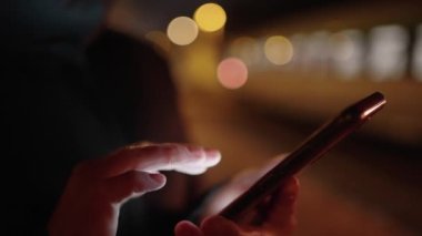 Şehir treninin platformunda siyah ceketli bir kadın toplu taşıma durağında beklerken akıllı telefondan mesaj uygulaması kullanıyor.