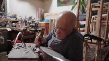 Senior elderly master jeweler in wheelchair works in a workshop with an anvil