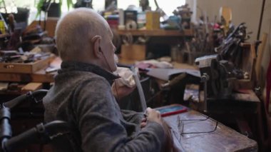 Senior gray haired male master in a wheelchair is working at his workshop sharpening japanese swords blade