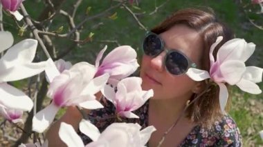 Güneş gözlüklü mutlu kadın doğanın tadını çıkarıyor ve bahçede çiçek açan manolya çiçeklerine dokunuyor.
