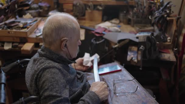 車椅子のシニアグレーの髪の男性マスターは 日本刀の刃を研ぐ彼のワークショップで働いています — ストック動画