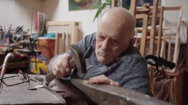Senior elderly master jeweler in wheelchair works in a workshop with an anvil