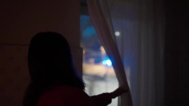 Genç bir kadın evinin penceresinden dışarı bakıyor. Odaklanamayan polis ışıklarıyla olay yerini inceliyor.