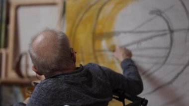 Tekerlekli sandalyedeki kıdemli ressam bir stüdyoda tuvale yağlı boya bir resim çizer.