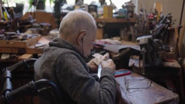 Senior gray haired male master in a wheelchair is working at his workshop sharpening japanese swords blade