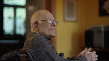 Tekerlekli sandalyede oturan yaşlı bir adam şöminenin yanında televizyon izliyor.