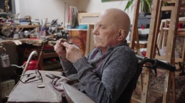 Senior elderly master jeweler in wheelchair works in a workshop with an anvil