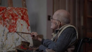 Tekerlekli sandalyedeki kıdemli ressam bir stüdyoda tuvale yağlı boya bir resim çizer.