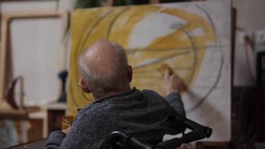 Tekerlekli sandalyedeki kıdemli ressam bir stüdyoda tuvale yağlı boya bir resim çizer.