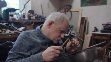 Tekerlekli sandalyede usta bir zanaatkar atölyesinde çalışıyor. Japon kılıcının ucuna çekiç ve keskiyle hormon resmi işliyor.