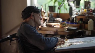 Tekerlekli sandalyedeki kıdemli sanatçı tuvaldeki yağlı boyaya altın sürüyor.