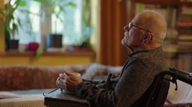 Tekerlekli sandalyede oturan yaşlı bir adam şöminenin yanında televizyon izliyor.