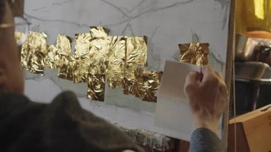 Tekerlekli sandalyedeki kıdemli sanatçı tuvaldeki yağlı boyaya altın sürüyor.