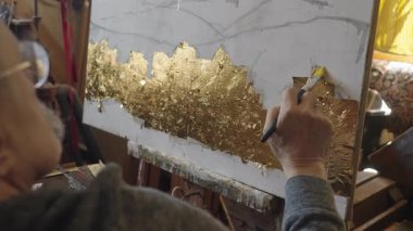 Tekerlekli sandalyedeki kıdemli sanatçı tuvaldeki yağlı boyaya altın sürüyor.