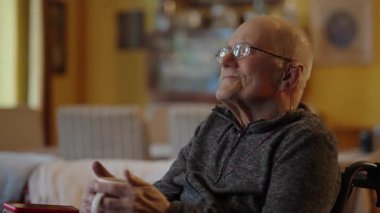 Tekerlekli sandalyede oturan yaşlı bir adam şöminenin yanında televizyon izliyor.