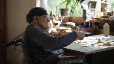 Tekerlekli sandalyedeki kıdemli sanatçı tuvaldeki yağlı boyaya altın sürüyor.