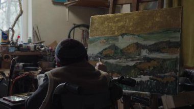 Tekerlekli sandalyedeki kıdemli ressam bir stüdyoda tuvale yağlı boya bir resim çizer.