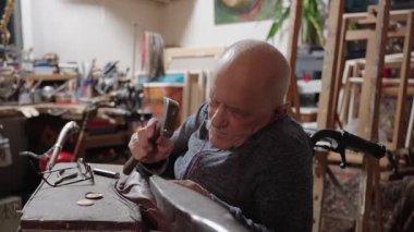 Senior elderly master jeweler in wheelchair works in a workshop with an anvil