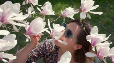 Güneş gözlüklü mutlu kadın doğanın tadını çıkarıyor ve bahçede çiçek açan manolya çiçeklerine dokunuyor.