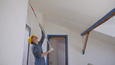 Yetişkin bir adam silindirle bir duvar boyuyor, tavan arası zemininde tamirat yapıyor.