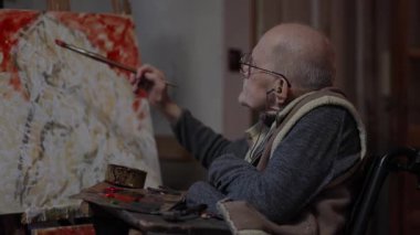 Tekerlekli sandalyedeki kıdemli ressam bir stüdyoda tuvale yağlı boya bir resim çizer.