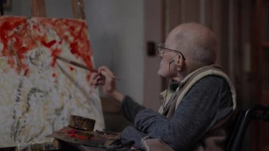 Tekerlekli sandalyedeki kıdemli ressam bir stüdyoda tuvale yağlı boya bir resim çizer.