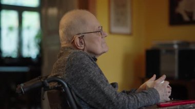 Tekerlekli sandalyede oturan yaşlı bir adam şöminenin yanında televizyon izliyor.