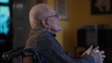 Tekerlekli sandalyede oturan yaşlı bir adam şöminenin yanında televizyon izliyor.