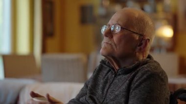 Tekerlekli sandalyede oturan yaşlı bir adam şöminenin yanında televizyon izliyor.