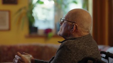 Tekerlekli sandalyede oturan yaşlı bir adam şöminenin yanında televizyon izliyor.