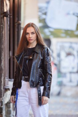 Şehirde poz veren deri ceketli ve kot pantolonlu güzel bir kız.