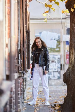 Siyah deri ceketli, gülümseyen güzel bir kadın şehirde yürüyor.