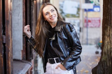 Siyah deri ceketli güzel, gülümseyen genç bir kadın sokakta dikiliyor.