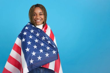 Amerikan bayrağına sarılmış Afrikalı bir kadının mavi arka planlı stüdyo portresi.
