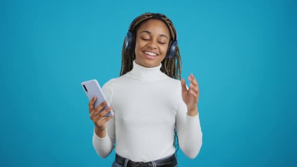 一个笑着的非洲女人一边看着相机一边用手机听音乐的蓝色背景视频 — 图库视频影像